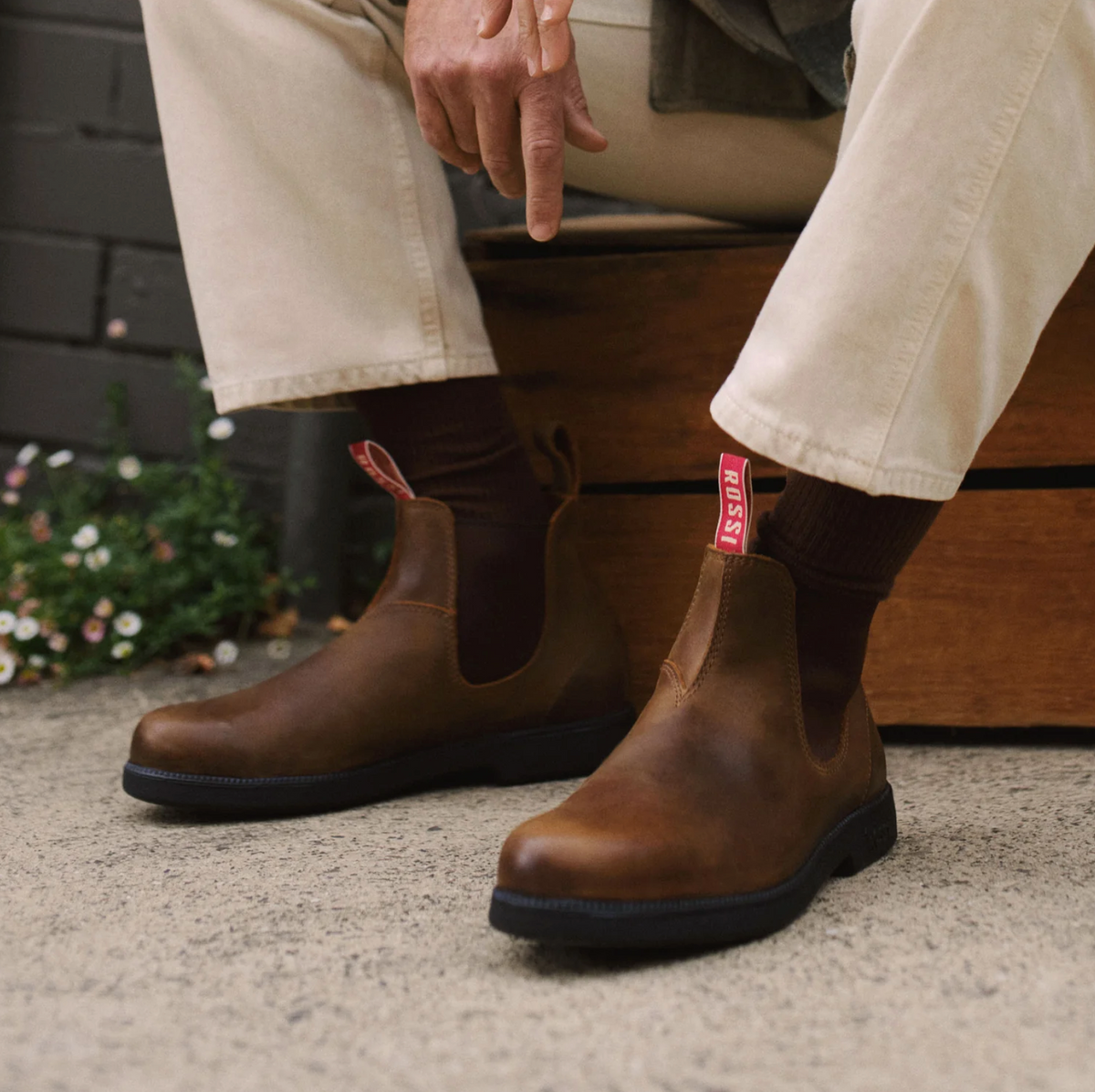 A person is seated, wearing light-colored pants and Rossi Booma 607 Elastic Sided Boots made from Full Grain Australian Leather, featuring distinctive red pull tabs. The boots are paired with dark socks. One hand rests on their knee, and a wooden box can be seen in the background with some greenery to the left.