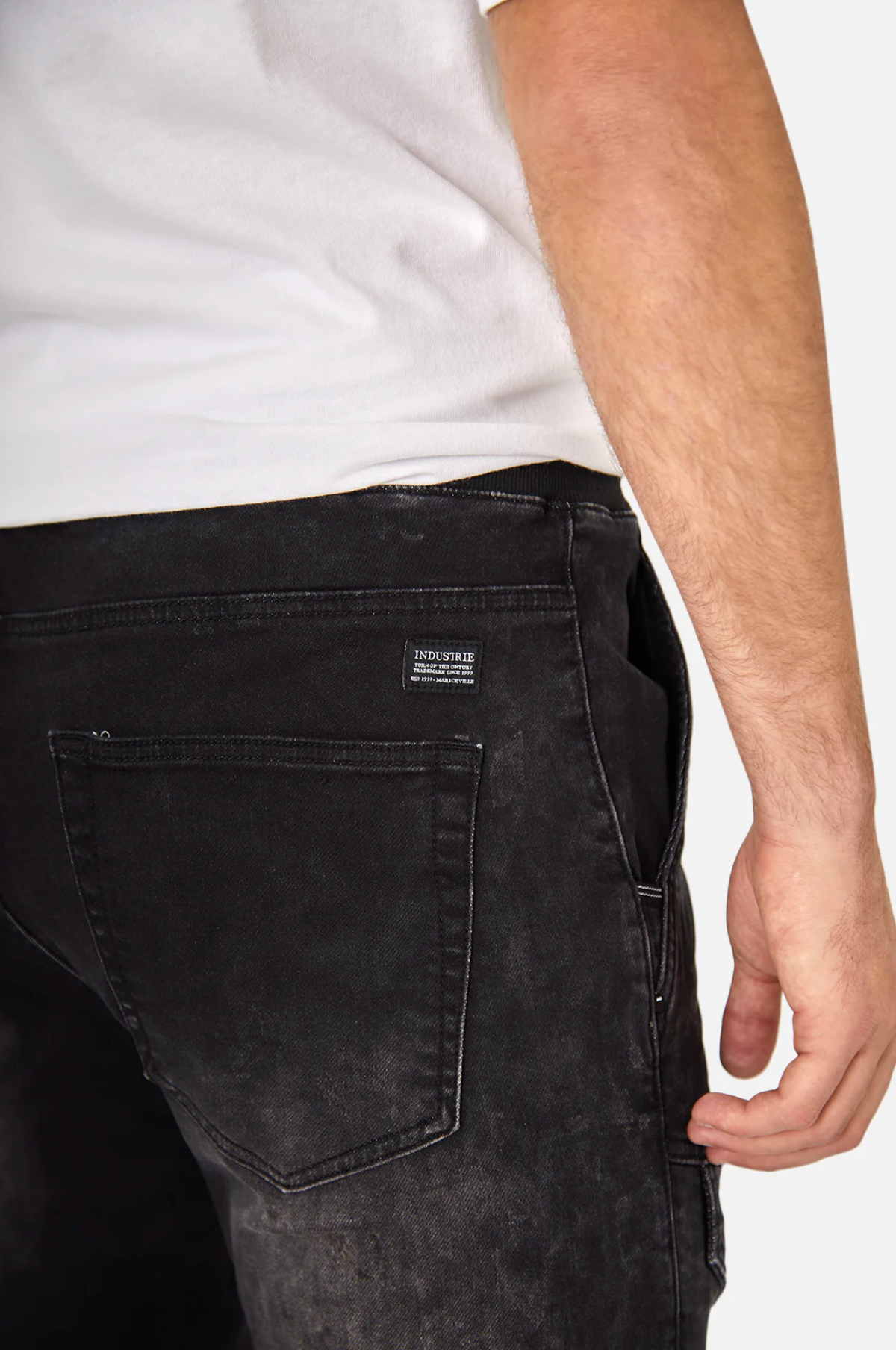 Close-up of a person wearing Industrie The Denim Armoured Drifter Pant with a white t-shirt. The black jeans showcase a small label above the back pocket and feature subtle knee panel detailing. Part of their right arm is visible.