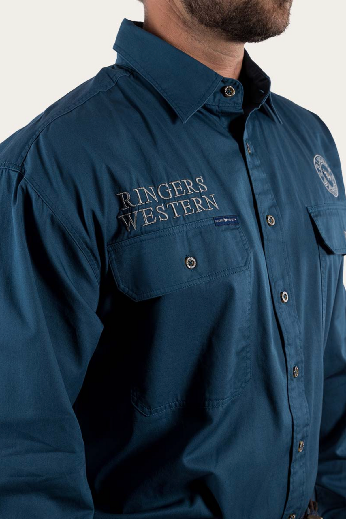 A man wears a dark blue Ringers Western Hawkeye Embroidered Shirt with "Ringers Western" on the chest, UPF50+ protection, two front pockets, and a partial logo on the left shoulder against a plain white background.