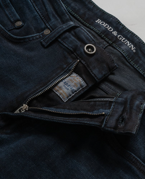 Close-up of Rodd & Gunn Weston Straight Italian Denim Jeans with a partially open zipper and button clasp, an internal label visible. "Rodd & Gunn" is printed near the waistband on the deep blue-black fabric.