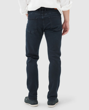 A person stands with their back to the camera, wearing Rodd & Gunn Weston Straight Italian Denim Jeans in deep blue-black and a white shirt, complemented by gray sneakers against a plain, light-colored background.
