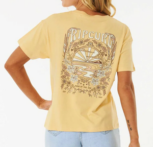 A woman wears the Rip Curl Riptide Relaxed Tee in light yellow, made from regenerated cotton jersey with a relaxed fit. The tee features a bold back print of a sunset, waves, flowers, and leaves as she stands with one hand on her hip.