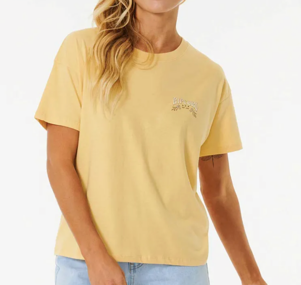 A person with long wavy blonde hair stands against a plain background, wearing the Rip Curl Riptide Relaxed Tee in pale yellow regenerated cotton jersey with a printed leaf and small left chest text, paired with light blue jeans.