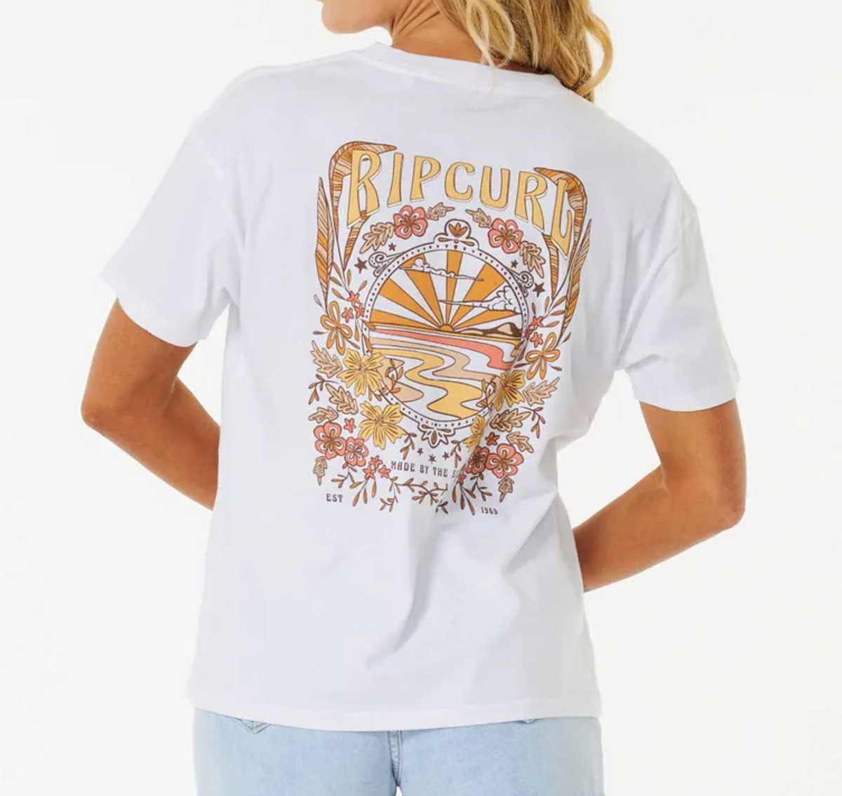A woman wears the Rip Curl Riptide Relaxed Tee in white, made from regenerated cotton jersey, featuring a colorful back print of a stylized sunset, waves, and flowers. She faces away with her hair draped over one shoulder.