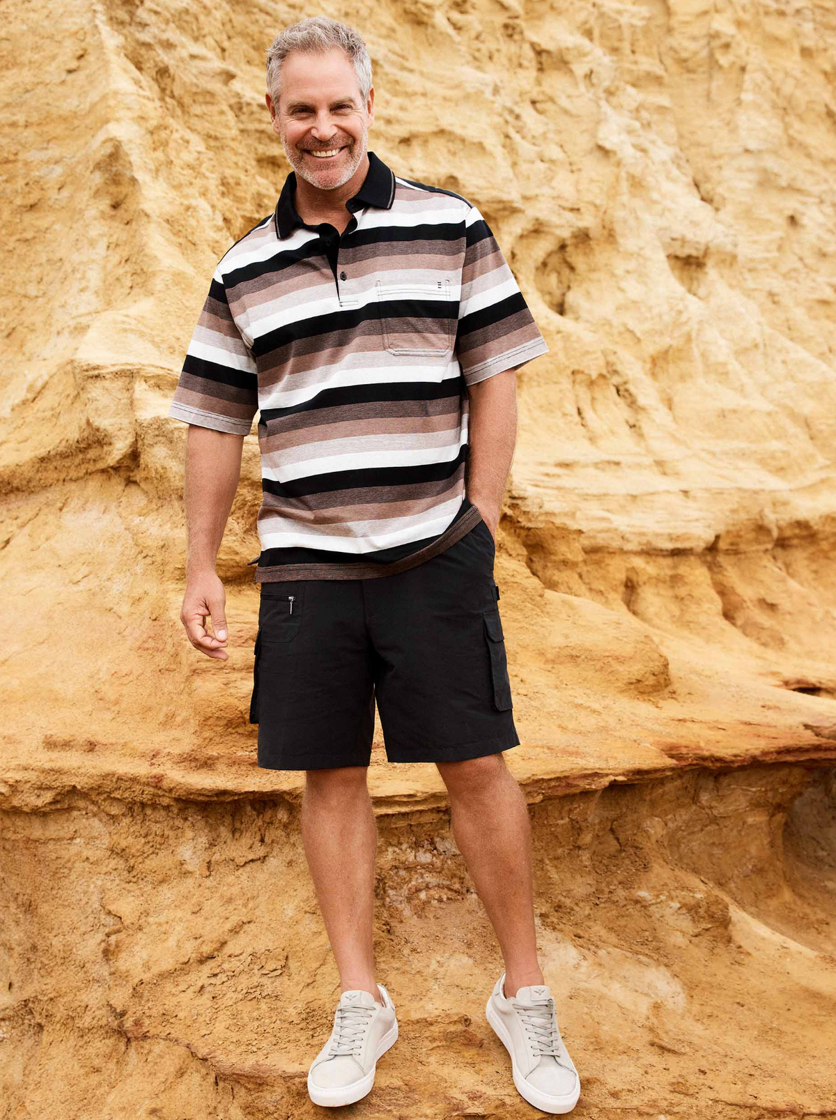 A smiling man with gray hair stands outdoors by a sandy, rocky cliff, dressed in the Breakaway Ked Mercerised Polo, black cargo shorts, and white sneakers.