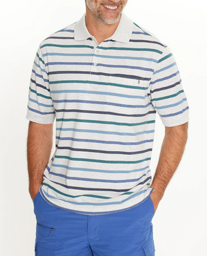Wearing a Breakaway Ozi Kinnersly Polo with a rib knit collar and white, gray, blue, green, and black accents, paired with bright blue pants, the person smiles warmly with hands in pockets.