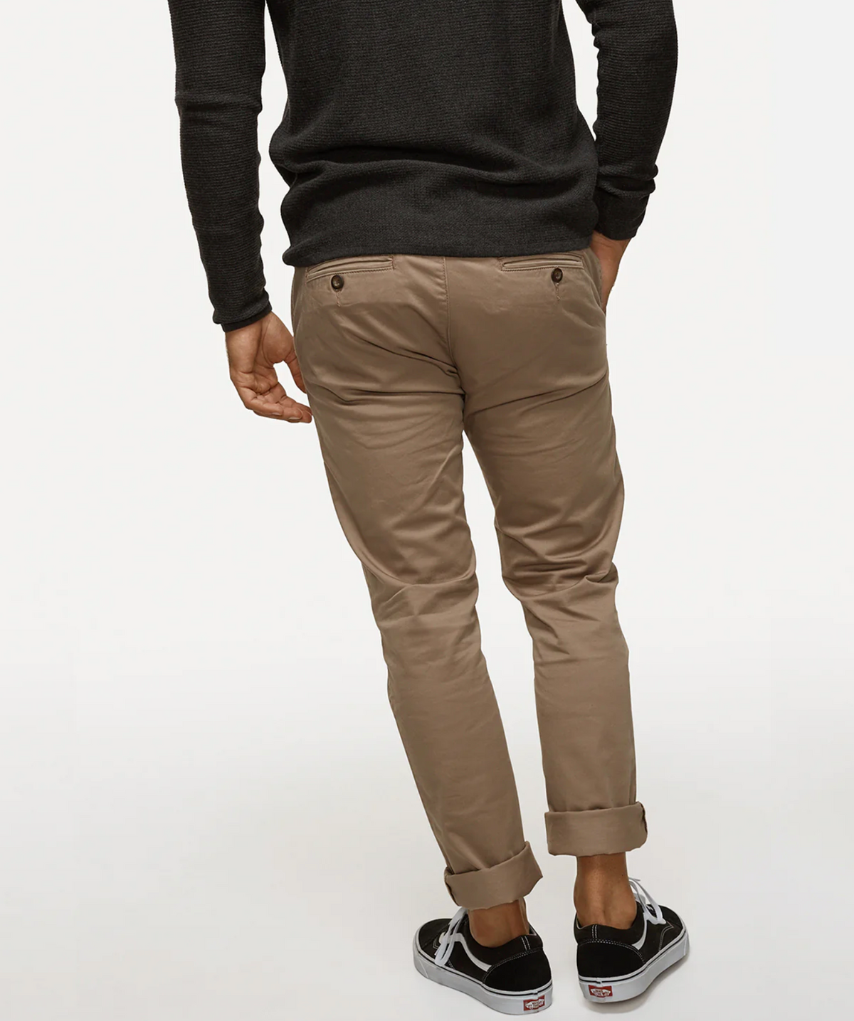 A person wearing a black sweater and beige Industrie The Regular Cuba Chino Pants stands with their back to the camera. The chinos, crafted from stretch cotton for extra comfort, feature two back pockets with buttons. They are also wearing black sneakers with white soles and heel tabs. The bottom edges of the wider leg fit chinos are rolled up.