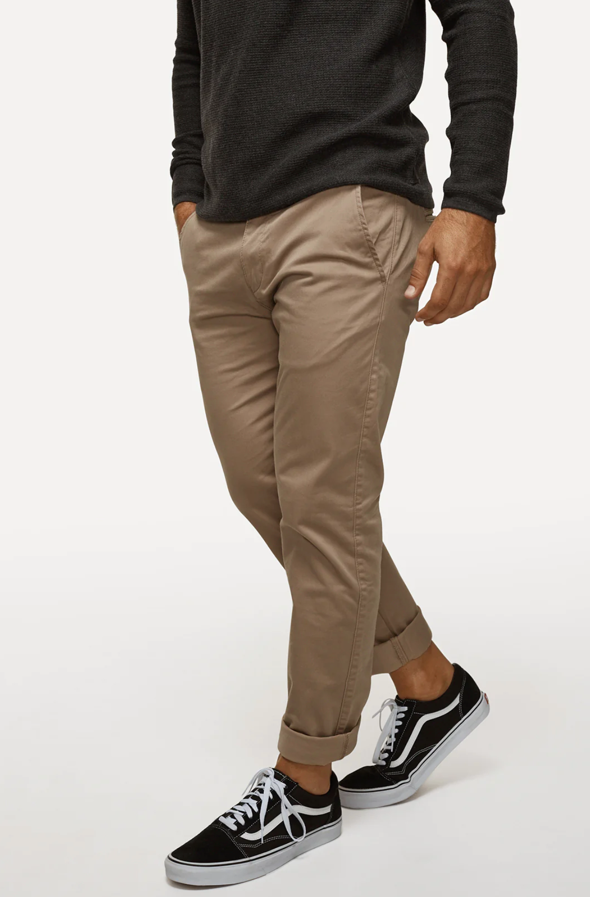 A person stands against a plain background wearing a long-sleeve, dark gray shirt, Industrie The Regular Cuba Chino Pant in beige with extra storage, and black and white sneakers. The person's left hand is in their pocket, and their right hand is relaxed by their side.