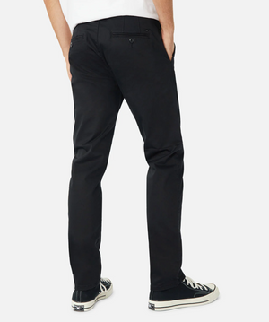 Back view of a person wearing Industrie The Regular Cuba Chino Pant in black, featuring a wider leg fit and paired with black sneakers with white soles. The person has their hands in their pockets. These durable stretch cotton pants include two back pockets with button closures, a small logo tag near the right pocket, and additional storage options.