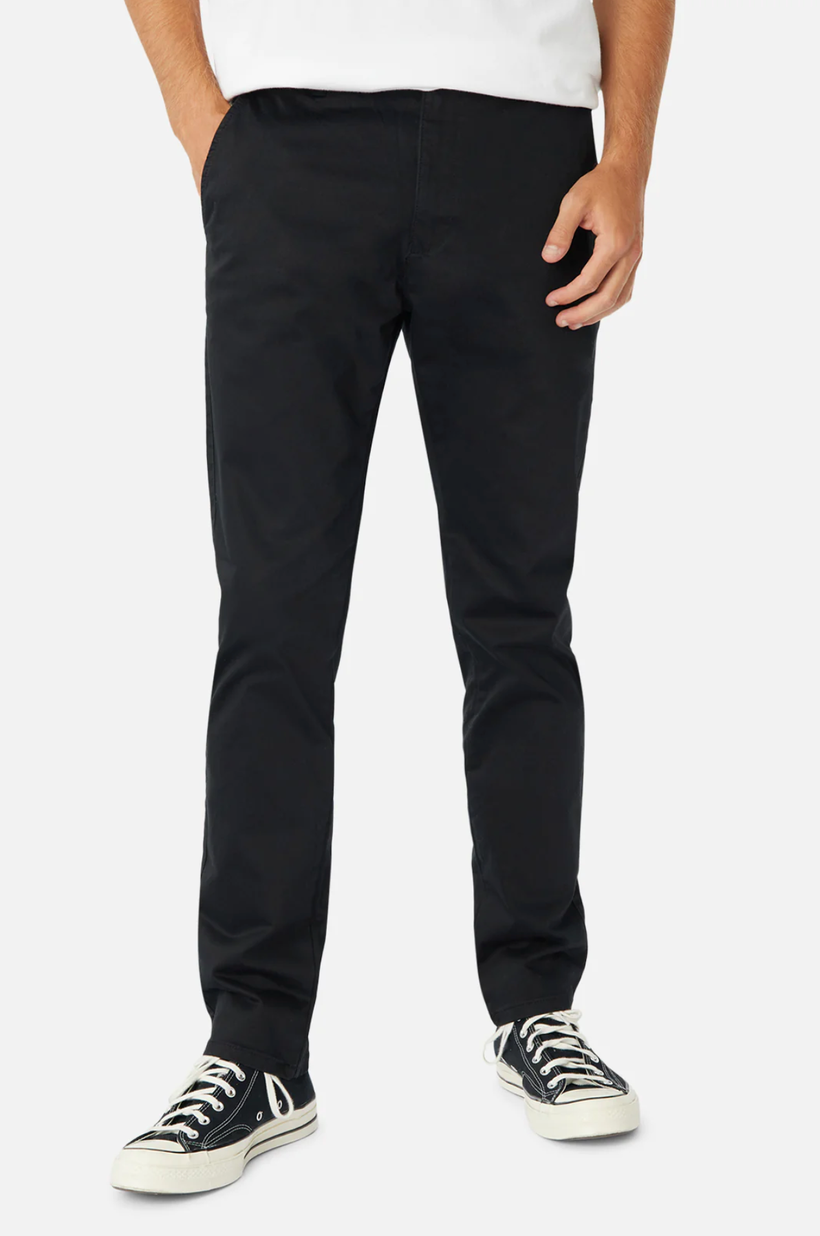 A person is wearing black Industrie The Regular Cuba Chino Pants made from durable stretch cotton and black-and-white sneakers, standing against a white background. Only the torso and legs are visible, with the person's left hand resting in the pocket of the chinos.