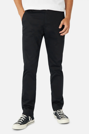A person is wearing black Industrie The Regular Cuba Chino Pants made from durable stretch cotton and black-and-white sneakers, standing against a white background. Only the torso and legs are visible, with the person's left hand resting in the pocket of the chinos.