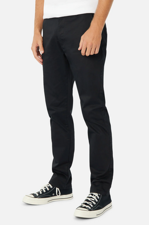 A person is standing against a plain white background, wearing Industrie The Regular Cuba Chino Pant, which features a wider leg fit. They are also wearing black canvas sneakers with white soles and laces. Only the lower half of the person's body is visible. The person is also wearing a white top.