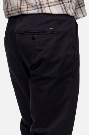 A close-up of the backside of a person wearing Industrie The Regular Cuba Chino Pant in a dark color and a light-colored plaid shirt. The durable stretch cotton pants feature a buttoned back pocket, side pockets, and are slightly crumpled at the back. The background is a plain, light-colored surface.