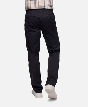 The image shows the back view of a person wearing the Industrie The Regular Cuba Chino Pant, which is dark-colored and made from durable stretch cotton, featuring side and back pockets. Paired with white sneakers, the ensemble is completed with a checkered shirt whose tail is partially visible against the plain white background.