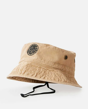 The Rip Curl Wetsuit Icon Mid Brim Hat is tan with a black chin strap, two metal eyelets per side, and a circular black patch with the Wetsuit Icon logo on the front. It’s shown against a white background.