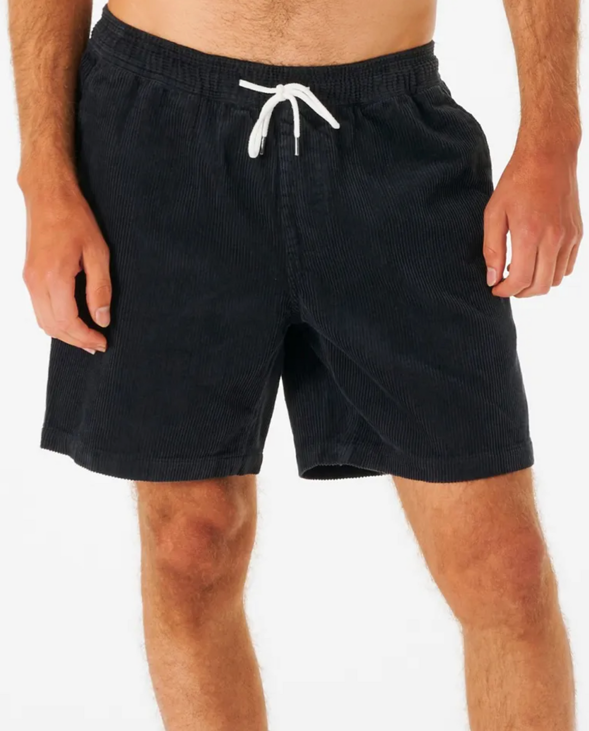 Wearing Rip Curl Surf Revival Cord Volley shorts with a white drawstring, a person stands against a plain backdrop. Only their lower torso and legs in the baggy fit black corduroy shorts are visible, evoking 90's style charm.