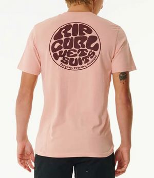 A person faces away, wearing the Rip Curl Wetsuit Icon Tee in light pink—a 100% cotton casual shirt with a large circular "Rip Curl Wetsuits" logo on the back and small text featuring Torquay, Trestles, and Hossegor.