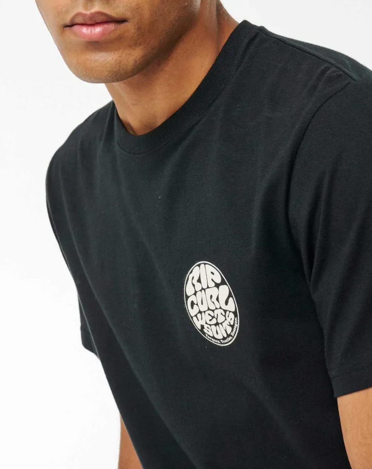 A person wears the Rip Curl Wetsuit Icon Tee, a black 100% cotton shirt with a small circular white logo reading "Rip Curl West Surf" on the left chest. The upper torso and part of the face are shown against a plain background.