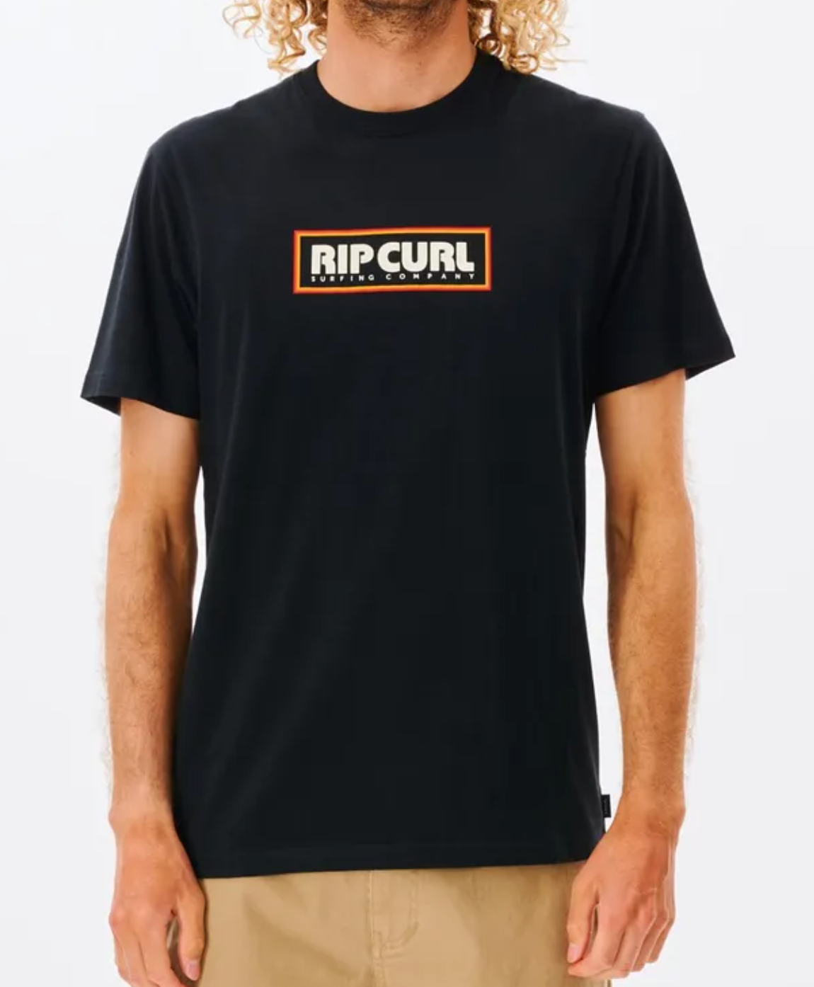 A person with curly hair wears a Rip Curl Big Mumma Icon Tee in organic cotton, showing "RIP CURL" in bold white letters inside an orange rectangle, with "SURFING COMPANY" beneath. They pair it with beige shorts against a plain white background.