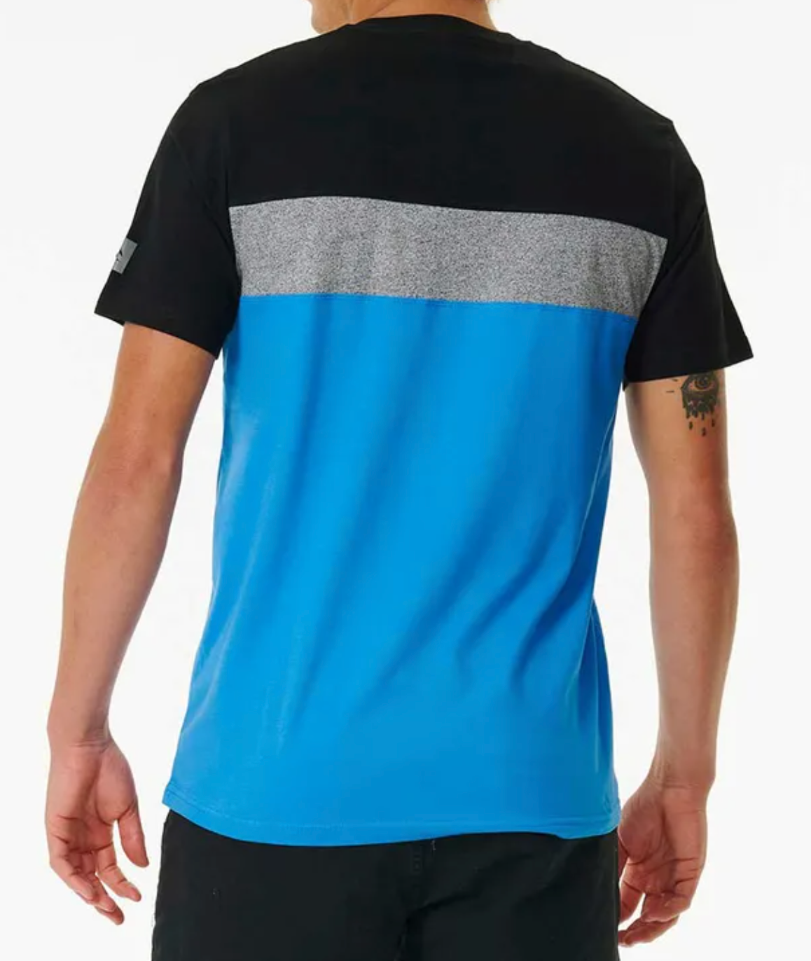 A person with short hair and a left arm tattoo is seen from behind wearing the Rip Curl Undertow Panel Tee, made of 100% cotton, featuring a black upper section, gray back stripe, and bright blue lower half.