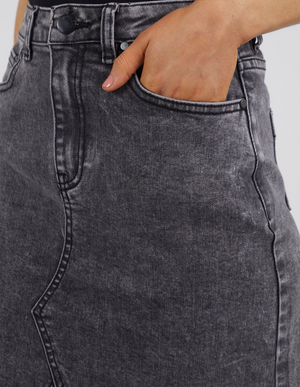 Wearing the Foxwood Belle Skirt, a high-waisted black denim piece with visible seams and a slightly faded texture, an individual effortlessly places their left hand inside the front pocket. The skirt’s A-line fit ensures both style and comfort.
