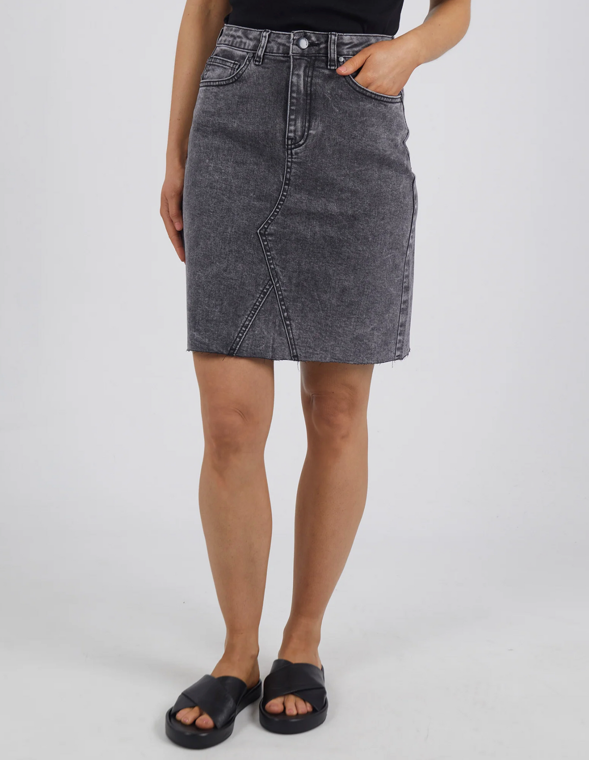 Against a plain white background, someone wears a black top with the Foxwood Belle Skirt in dark gray denim, featuring an A-line fit and diagonal seam detailing, and completes the look with black slide sandals.