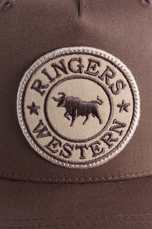 Close-up of the Ringers Western Signature Bull Trucker Cap, featuring a brown cotton twill patch with "RINGERS WESTERN" text, stars, and an embroidered bull in the center, stitched on a brown trucker cap with mesh back.
