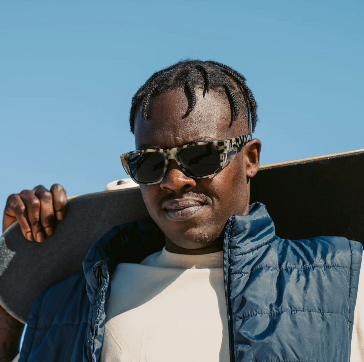 A person with short twists and a neutral expression holds a skateboard over their shoulder under a clear blue sky, wearing SafeStyle Fusions Milky Tort Frame sunglasses with polarised lenses and UV protection, paired with a blue vest.