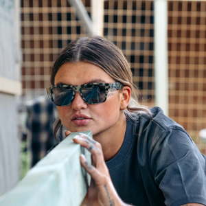 Wearing SafeStyle Fusions with a Milky Tort frame and polarized lenses, a person in a dark shirt thoughtfully examines a wooden beam. The blurred, grid-like backdrop suggests a construction site where UV protection and safety are crucial.