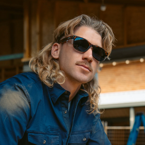 A man with wavy, shoulder-length blonde hair and a mustache sports SafeStyle Classics sunglasses with brown tort frames and polarized lenses. He pairs them with a blue button-up shirt, standing outdoors against a rustic wooden structure.