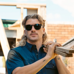 A man with wavy hair and a mustache wears SafeStyle Cruisers Brown Tort Frame Polarised Lens sunglasses and a navy button-up shirt, carrying wooden planks on his shoulder at a construction site with a fence and building in the background.