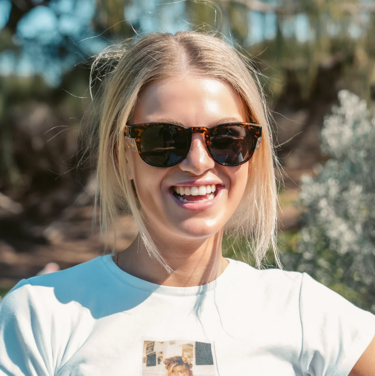 A young woman with blonde hair, wearing SafeStyle Cruisers Brown Tort Frame Polarised Lens sunglasses and a white T-shirt, smiles outdoors in bright sunlight with greenery softly blurred in the background.
