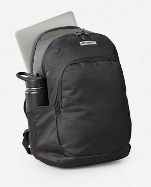 The Rip Curl Ozone Icons 30L Backpack in black holds a laptop in its back compartment and a water bottle in the side pocket. Made from recycled polyester, it features a top brand label. The sleek design stands out against the plain, light-colored background.