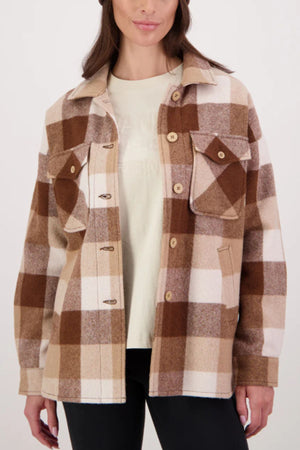 A woman wearing the Swanndri Anchorage V3 Women's Wool Shacket in beige and brown plaid over a light t-shirt and black pants stands against a plain background.