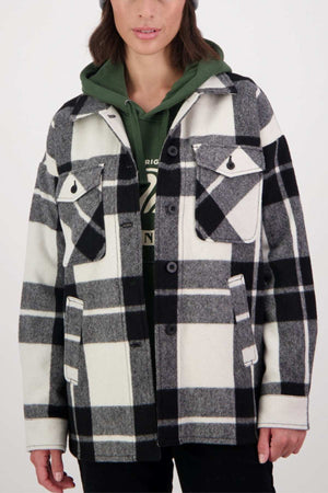 A person is wearing a black and white plaid Swanndri Anchorage v2 Wool Shirt Jacket over a green hoodie. The jacket, made from a blend of wool and merino, features two front pockets with flaps and black buttons down the front. The person has medium-length hair and is standing against a plain white background.