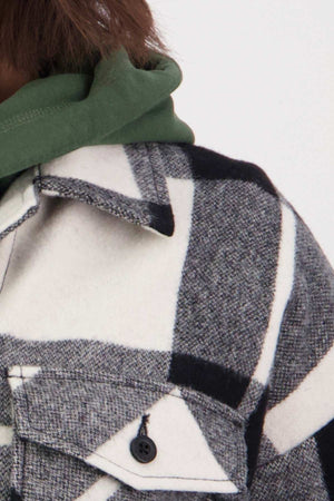 Close-up of a person wearing a green hoodie underneath a Swanndri Anchorage v2 Wool Shirt Jacket in black, white, and gray plaid. The coat features drop shoulders and welt hip pockets with large plaid patterns, while the green hood peeks over the collar.