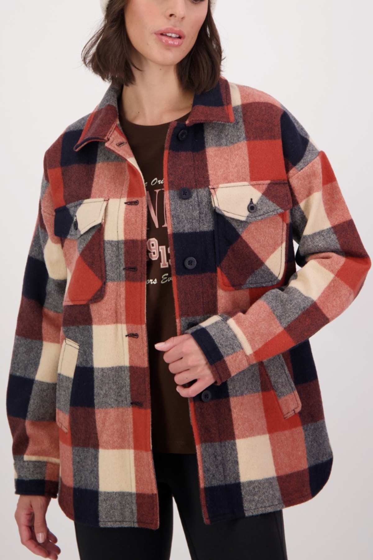A person in a Swanndri Anchorage v2 Wool Shirt Jacket with red, blue, and beige plaid over a brown shirt stands against a plain background. The jacket has two chest pockets and dark buttons for a timeless appearance.