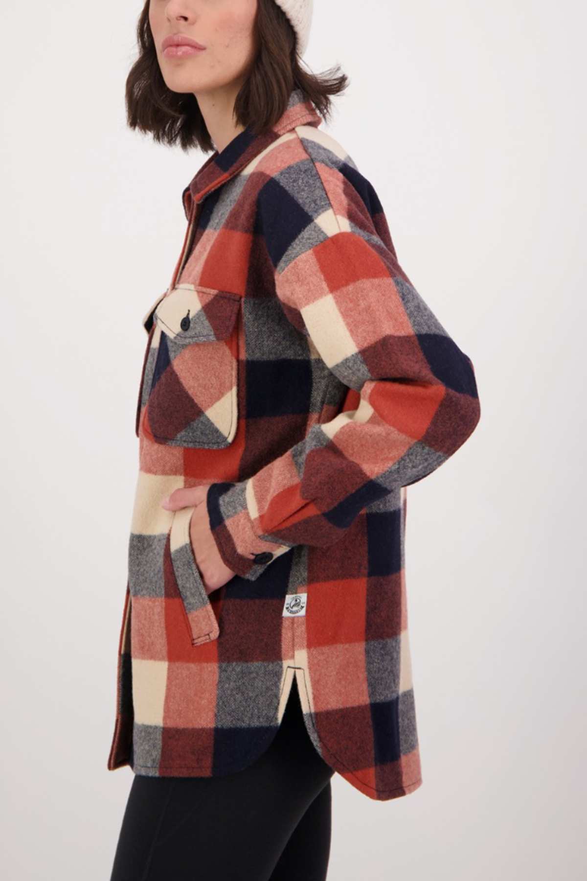 A person wears a Swanndri Anchorage v2 Wool Shirt Jacket with black pants and a beige beanie. The plaid jacket has a large chest pocket, and they are shown from the side, head slightly turned away.