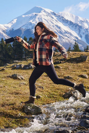 Leaping over a small stream, a person wears a Swanndri Anchorage v2 Wool Shirt Jacket, black leggings, and boots while holding a camera. Snow-capped mountains and pine trees appear in the sunny background.