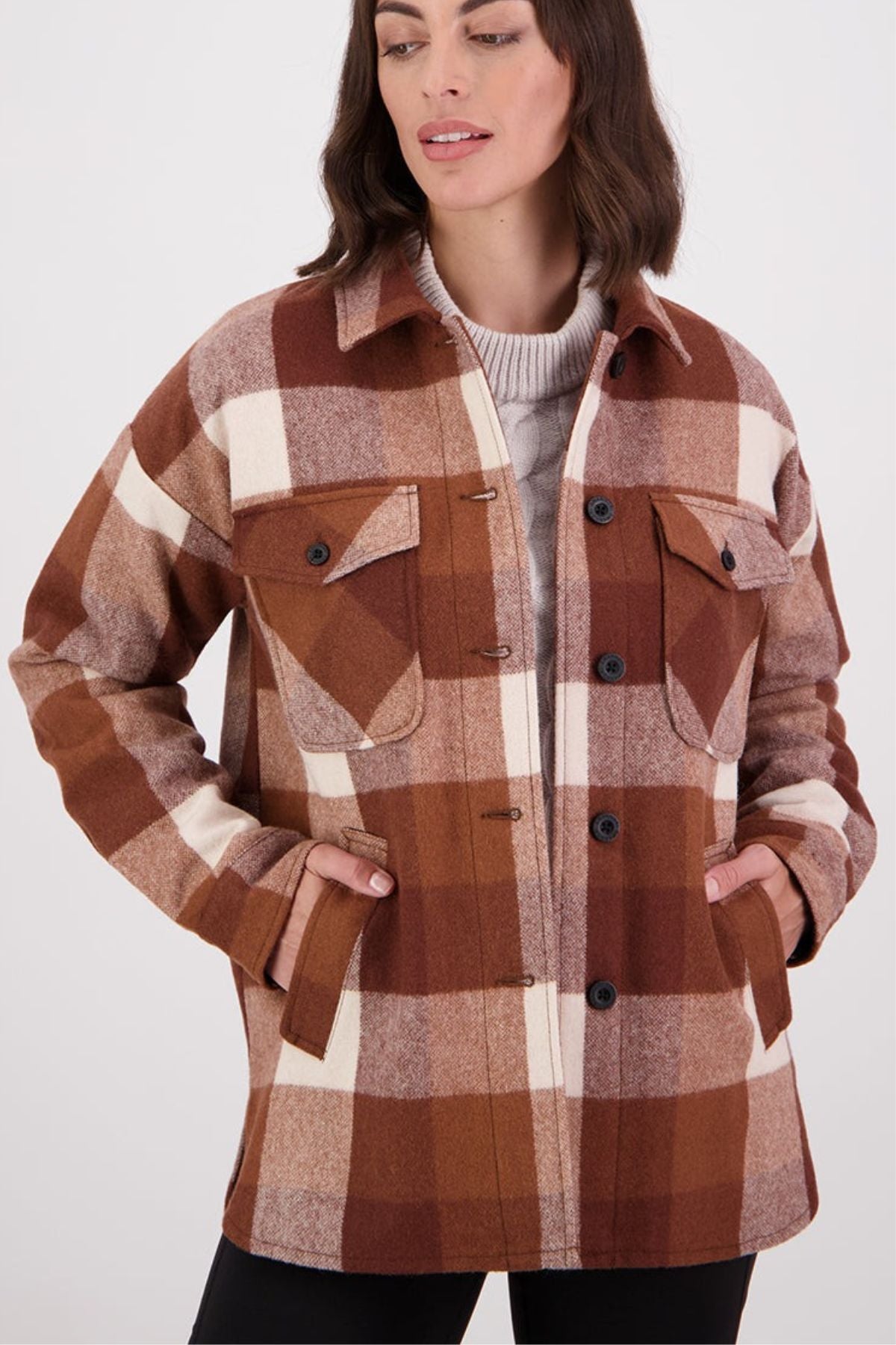 A woman wears the Swanndri Anchorage v2 Wool Shirt Jacket, a plaid button-up with front pockets, layered over a light beige sweater. Hands in her pockets, she looks slightly to the side and smiles softly.