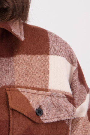 Close-up of a Swanndri Anchorage v2 Wool Shirt Jacket in brown, beige, and white plaid with a black button on the chest pocket; part of a person's shoulder and hair is visible.
