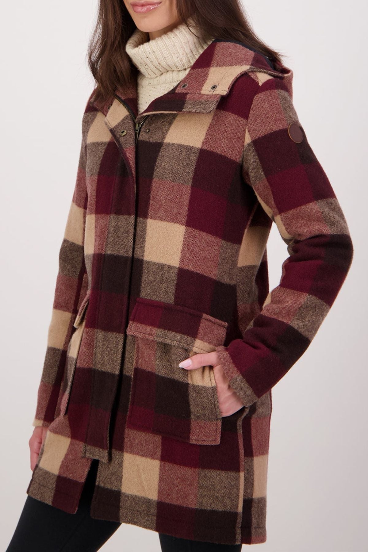 A woman wears a hooded, buttoned Swanndri Annmore V2 Wool Overcoat in large red, beige, and brown plaid over a cream turtleneck sweater, with her left hand in the coat pocket.