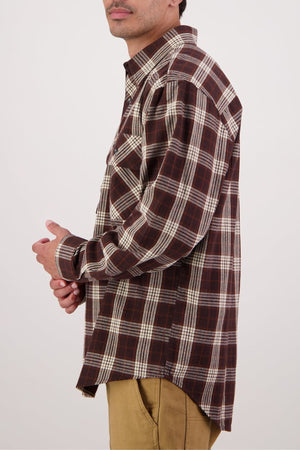A man in a brown and white plaid Swanndri Barn Shirt and tan pants stands sideways against a plain background, his face partly visible and hands clasped, showcasing the effortless heritage style of the Swanndri Barn Shirt.