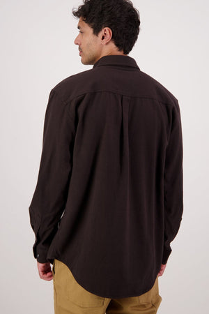 A man with dark hair, seen from behind, wears a Swanndri Barn Shirt in dark brown and tan pants, ideal for winter layering, against a plain light background.