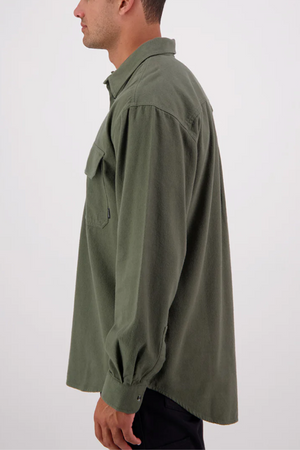 A person stands sideways in a Swanndri Barn Shirt—olive green, 100% cotton, chest pocket, relaxed fit—paired with black pants against a plain light background.