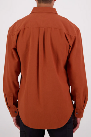 A person stands facing away, wearing a rust-colored Swanndri Barn Shirt in a relaxed fit made from 100% cotton, paired with black pants against a plain light background.