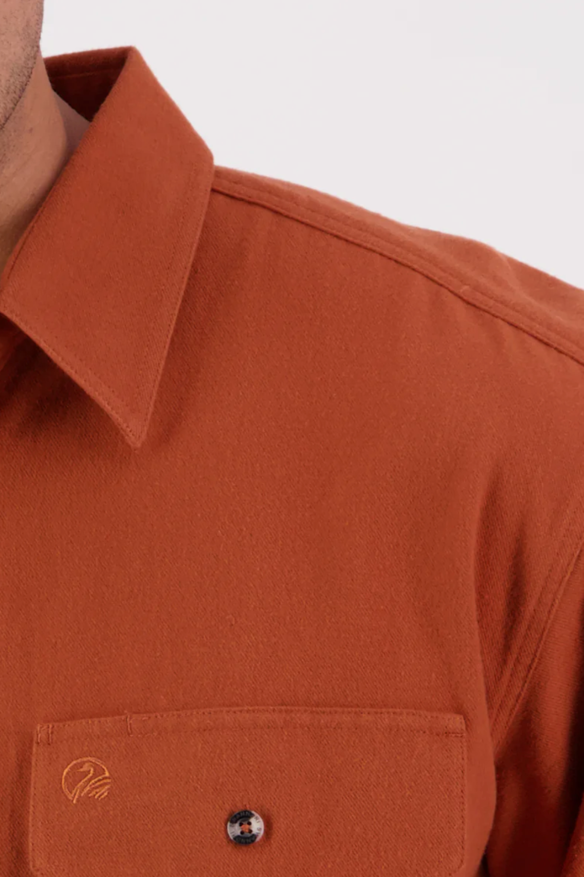 Close-up of the rust Swanndri Barn Shirt, highlighting the upper chest, collar, shoulder seam, and a buttoned pocket with embroidered detail. Made from 100% cotton with a relaxed fit. Shown against a plain light background.