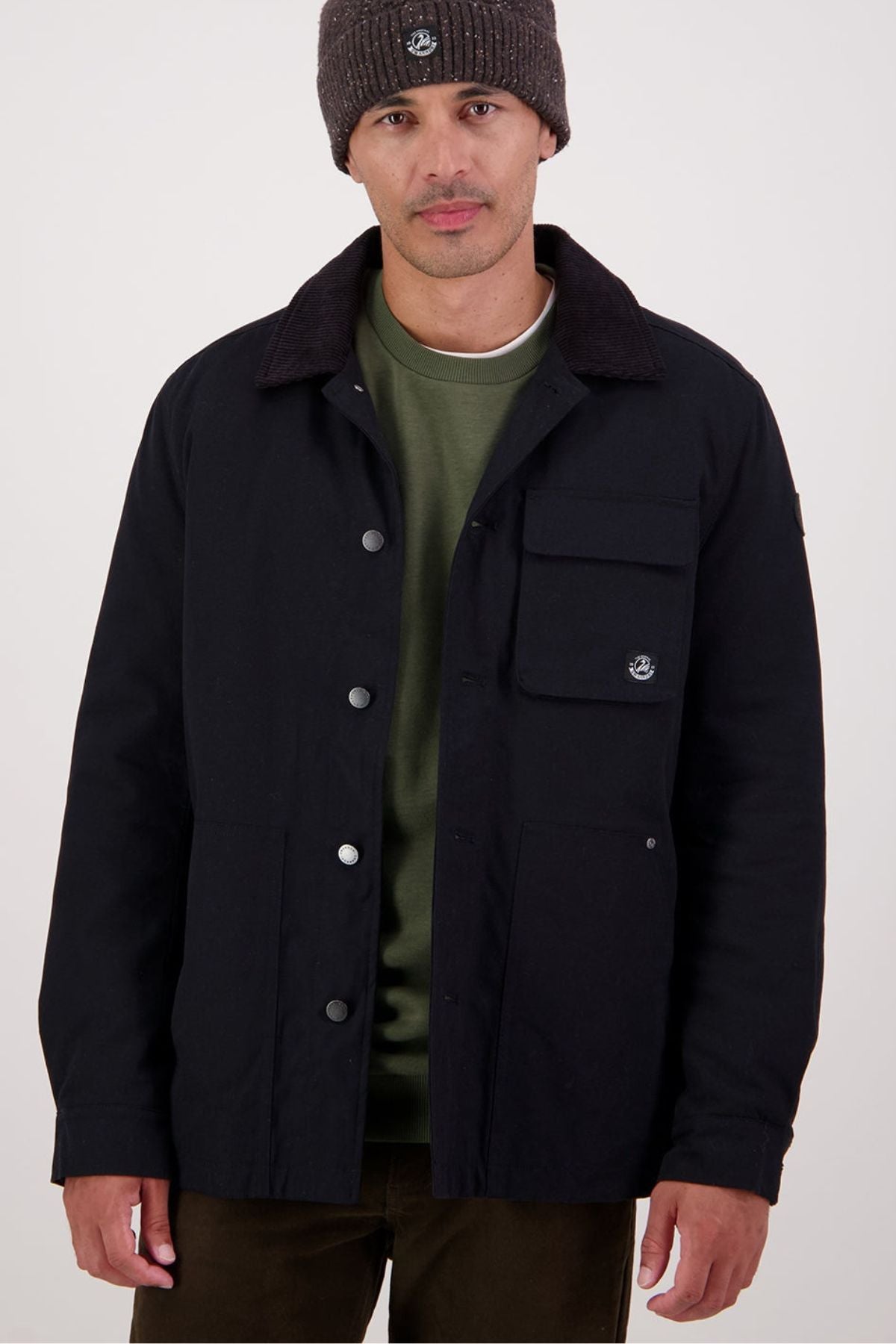 A man wearing a dark beanie, olive green sweatshirt, dark pants, and the Swanndri Barrytown V3 Canvas Jacket stands against a plain light background.