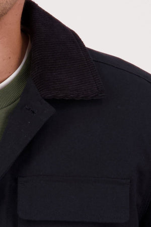 Close-up of a person wearing a black Swanndri Barrytown V3 Canvas Jacket with a black corduroy collar, layered over a green crewneck shirt. Only the lower half of their face and neck are visible.
