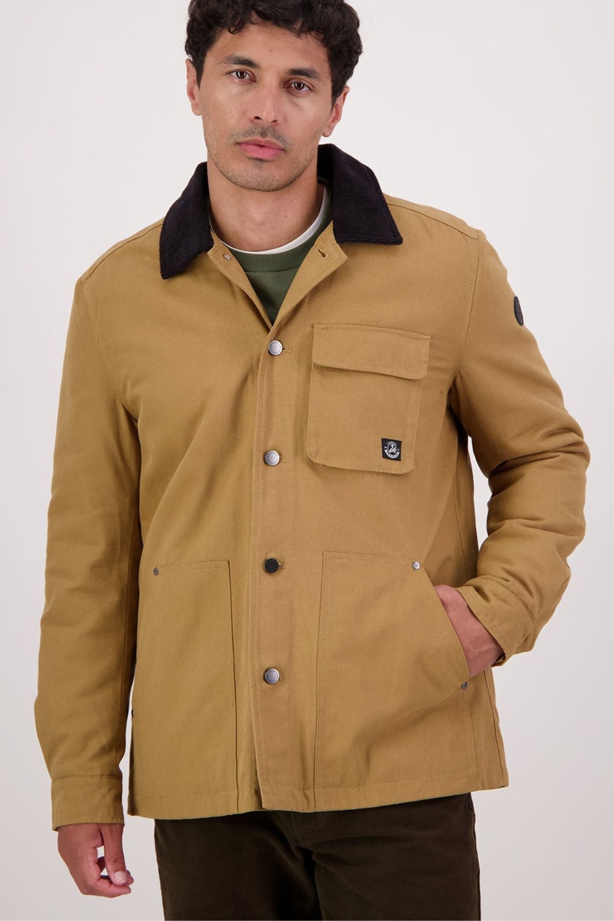 A man stands against a plain white background wearing the Swanndri Barrytown V3 Canvas Jacketโa tan cotton canvas button-up with large front pockets and a black collarโlooking at the camera with a neutral expression.